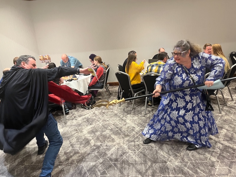 Two wandering NPCs fight a magical battle as tables of gamers play the UK Games Expo Epic